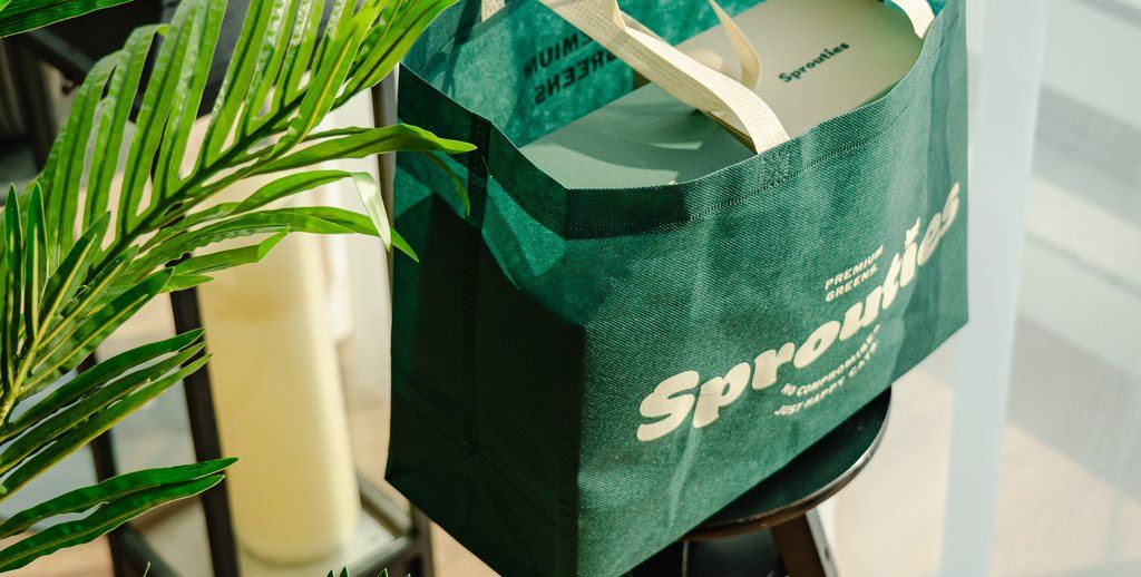 A green Sprouties tote bag holding a box of grass, on a stool among plants.
