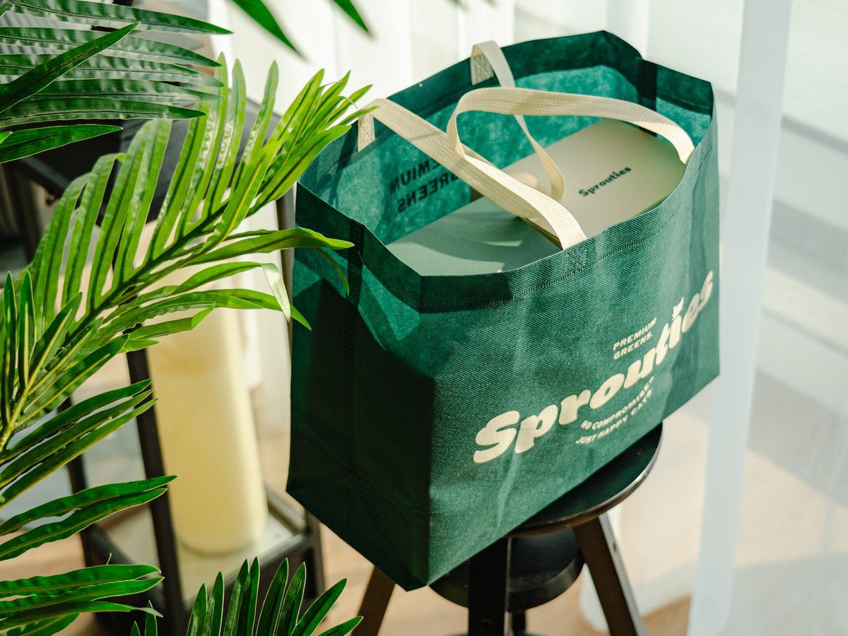 A green Sprouties tote bag holding a box of grass, on a stool among plants.